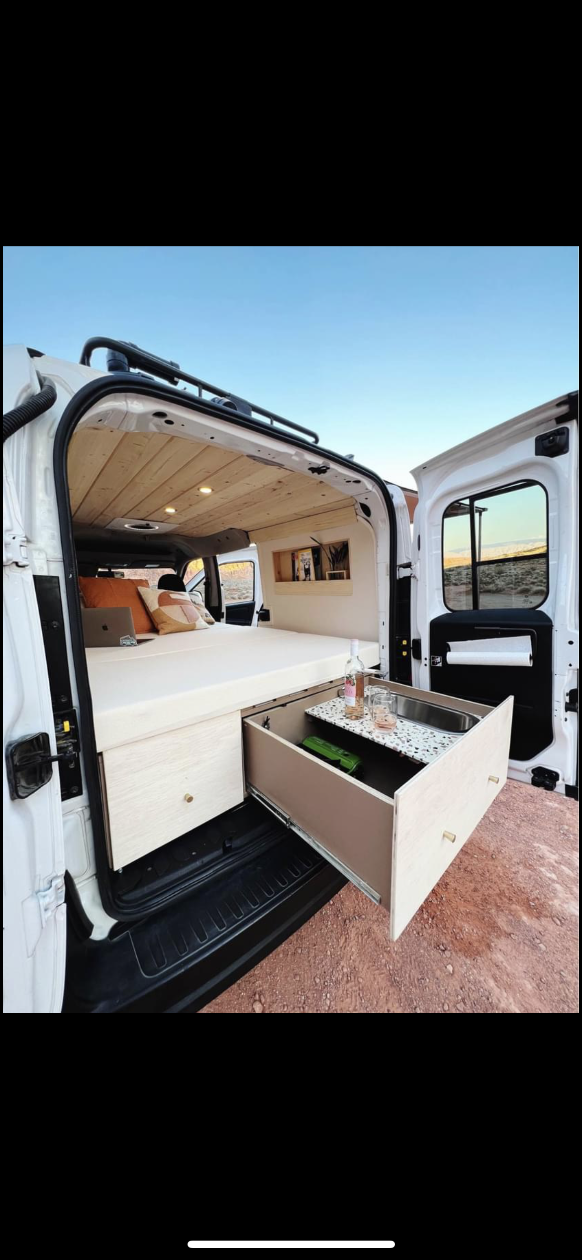 Stylish camper van RV rental interior: cozy bed, wood ceiling, and pull-out kitchenette visible in desert landscape.