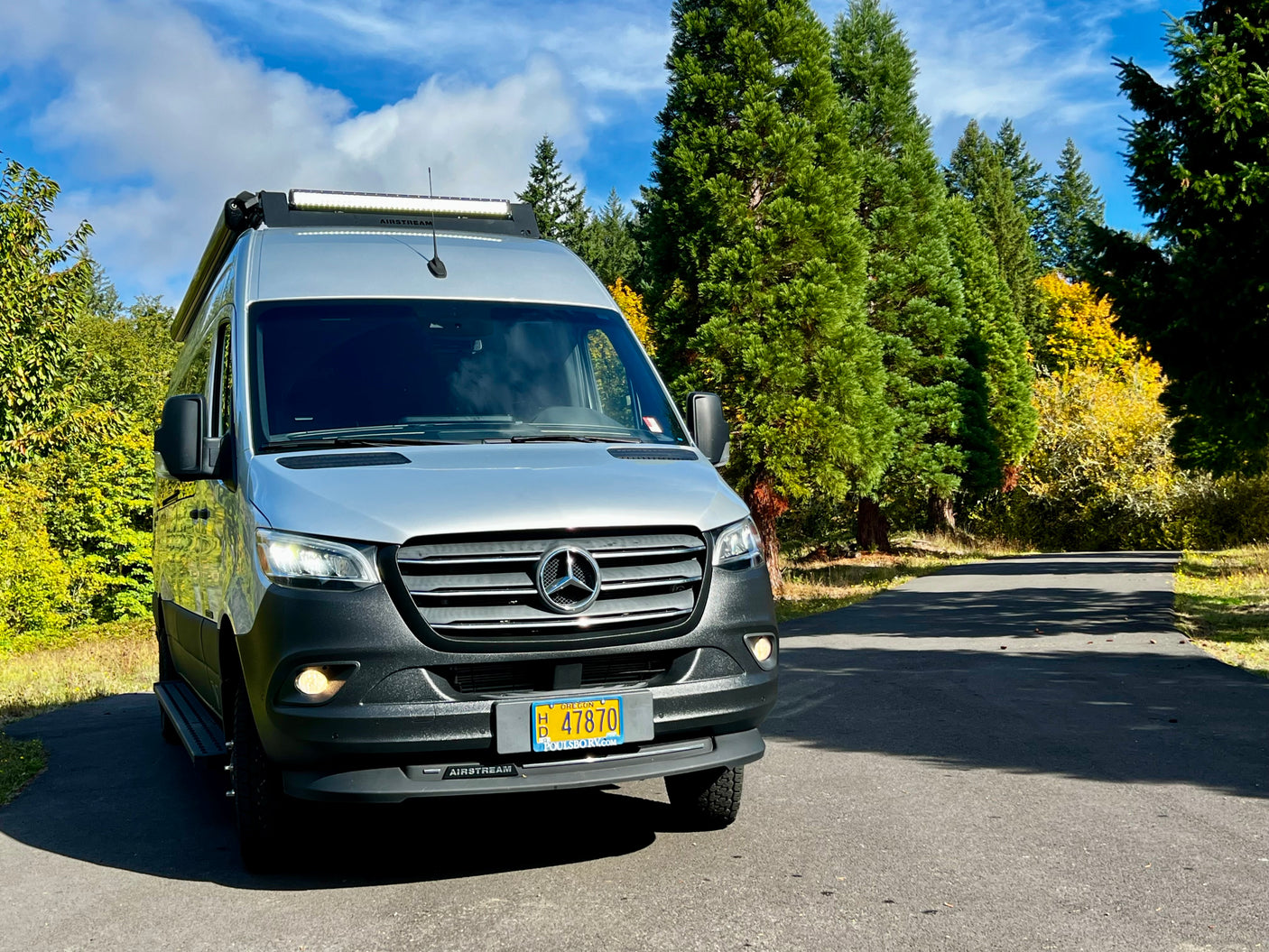 Silver Airstream Sprinter RV rental van parked on an asphalt road among green and yellow trees under a blue sky.