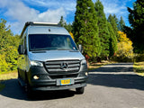 Silver Airstream Sprinter RV rental van parked on an asphalt road among green and yellow trees under a blue sky.