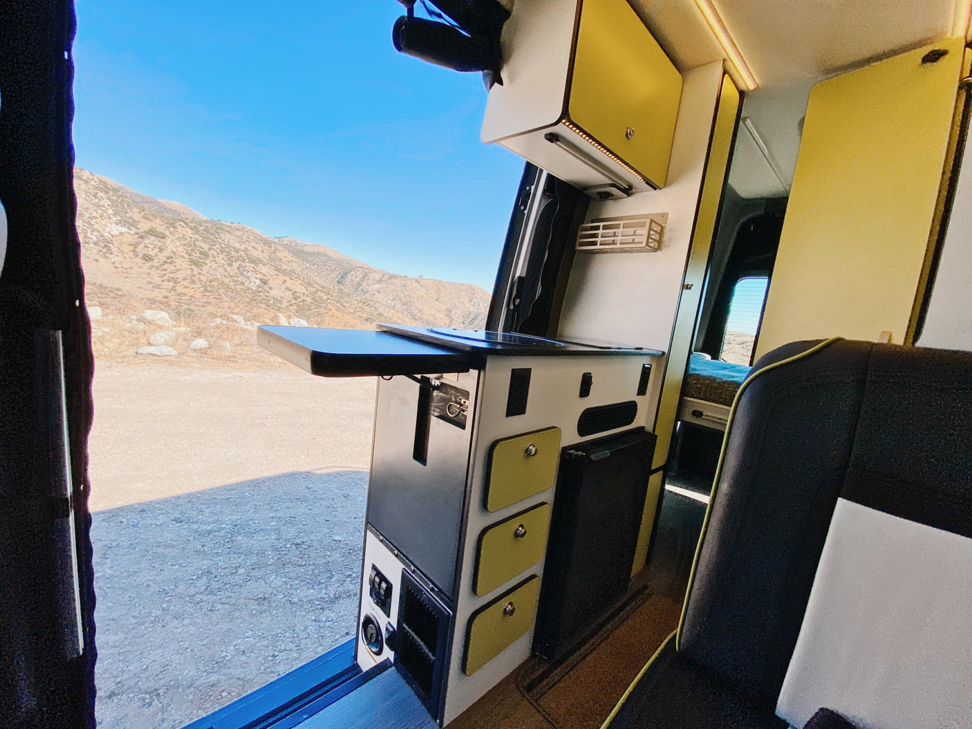 Inside an RV rental: a modern campervan kitchen with a pull-out table overlooks a scenic mountain landscape.