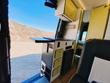 Inside an RV rental: a modern campervan kitchen with a pull-out table overlooks a scenic mountain landscape.