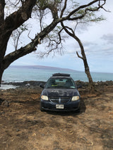 Dark minivan with roof carrier (an RV rental possibility) on Hawaii's rocky coast by the ocean, framed by trees.