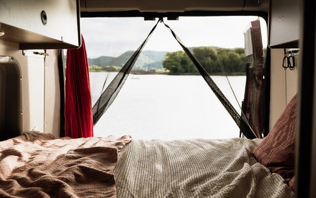 Inside an RV rental, a comfortable bed offers a relaxing view of a calm lake or river, complete with mosquito netting.