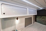 Stylish RV rental interior features white overhead cabinets, sleek LED lighting, a small wooden lamp, and a cozy bed by a window.