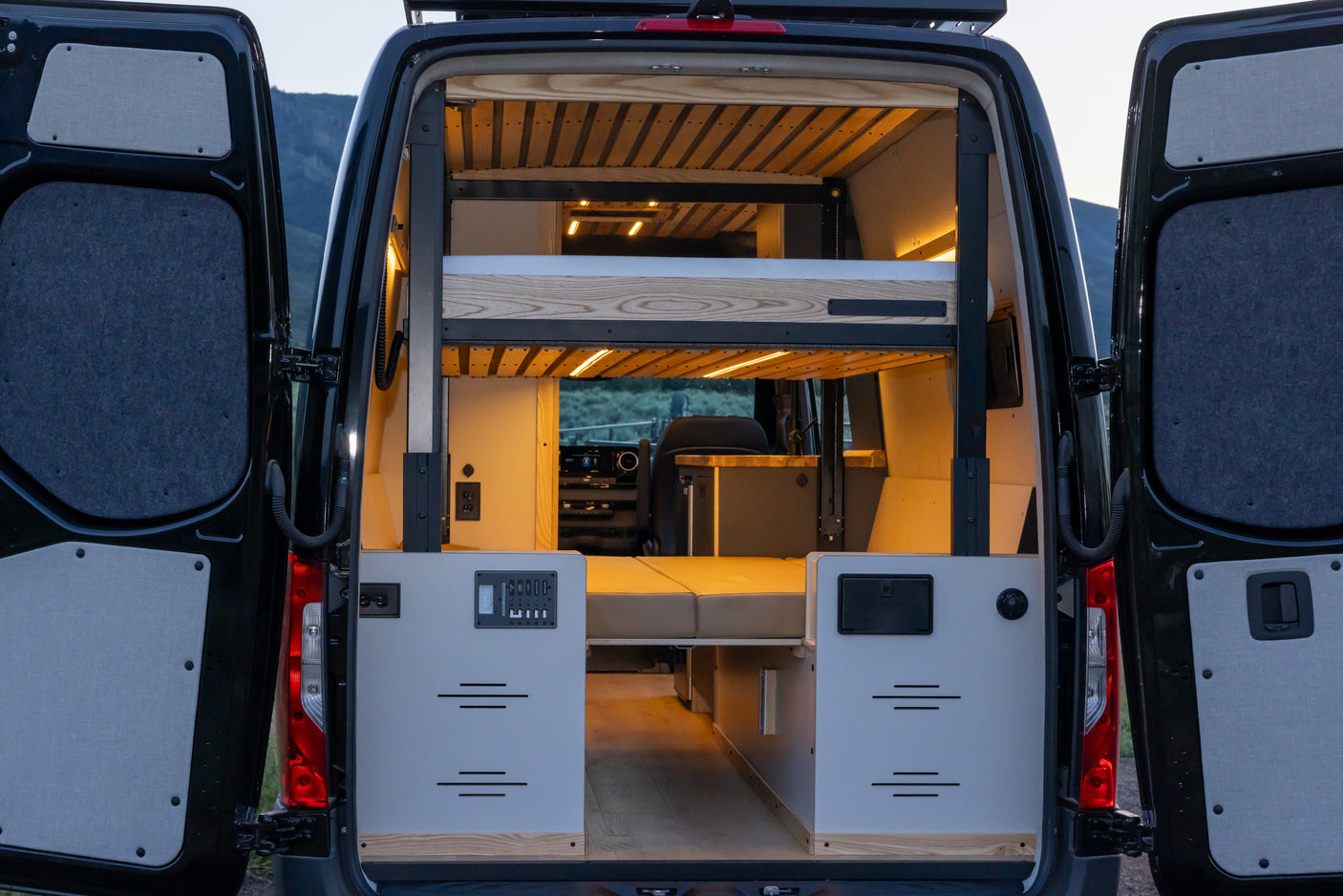 Opened rear doors of an RV rental camper van reveal a bright interior with wooden bunk beds, comfy cushions, and a control panel.