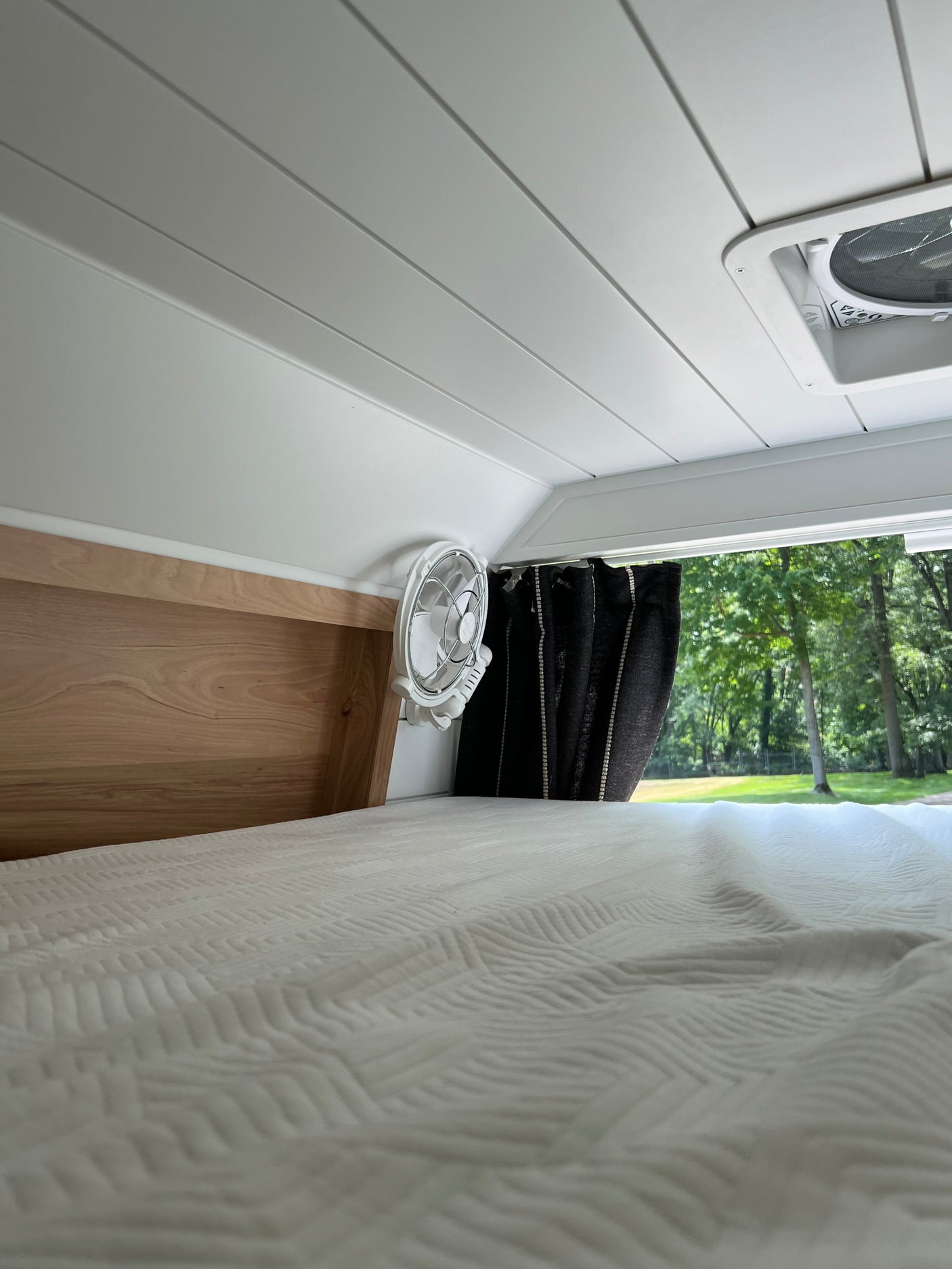 Inside a cozy RV rental: white bed, ceiling vent, small fan, and a serene outdoor view of green trees.