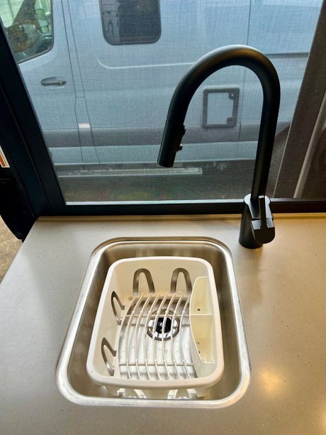 Close-up of an RV rental kitchen sink with a white dish rack, modern black faucet, and screened window view.