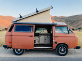 Scenic RV rental: Orange pop-top camper van with open door showing Westfalia kitchen. Mountains and cows in the background.