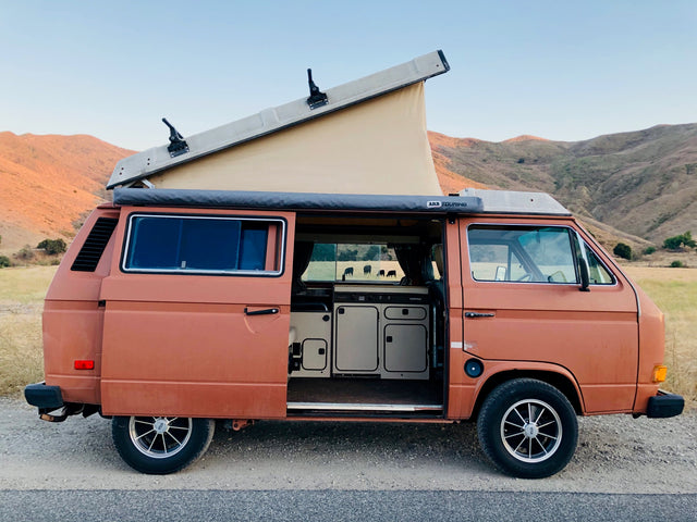 Scenic RV rental: Orange pop-top camper van with open door showing Westfalia kitchen. Mountains and cows in the background.