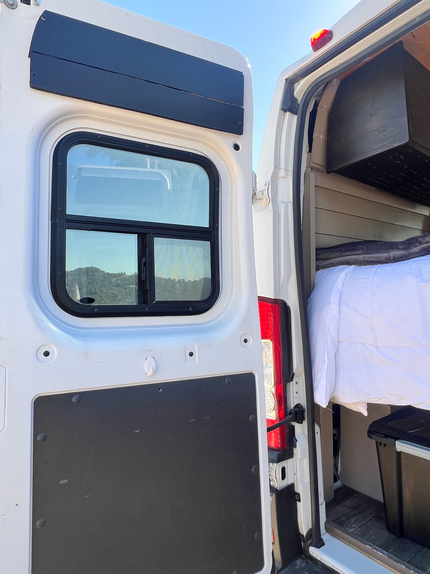White rv rental van's open rear doors reveal a cozy interior with a bed, wood panels, and a window showing mountain views.