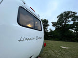 View of a white Happier Camper RV rental's rear, parked on grass at dusk, with interior lights glowing.
