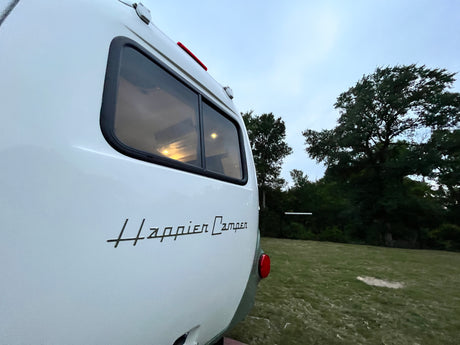 View of a white Happier Camper RV rental's rear, parked on grass at dusk, with interior lights glowing.