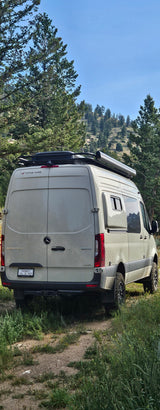 This light-colored Mercedes-Benz Sprinter camper van, possibly an RV rental, is parked on a dirt path in a scenic, treed setting.