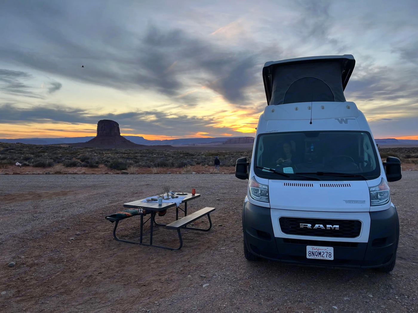  textured blankets & pillows.|Cozy RV rental pop-top bed overlooks saguaro cacti in a sunny desert scene.|This **RV rental**'s compact white wet bath features a toilet & integrated shower area with drain.|RV rental interior: A cream bench seat with two black seatbelts — photo 17
