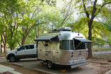 An Airstream RV rental, with awnings extended, is hitched to a white truck at a scenic, tree-filled campsite.