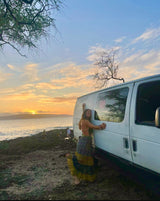 Woman embraces her white RV rental van at sunset by the ocean on a rocky shore, trees overhead. Perfect travel.