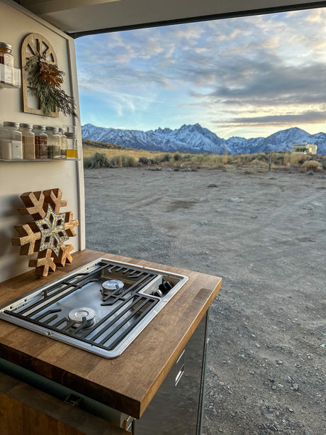 This RV rental offers a kitchen with a stove and spices, overlooking majestic snowy mountains, a vast camping area, and a distant RV.