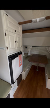 Bright RV rental interior: white cabinets, fridge with pink towel, and cozy dinette with wooden table and light green cushions.