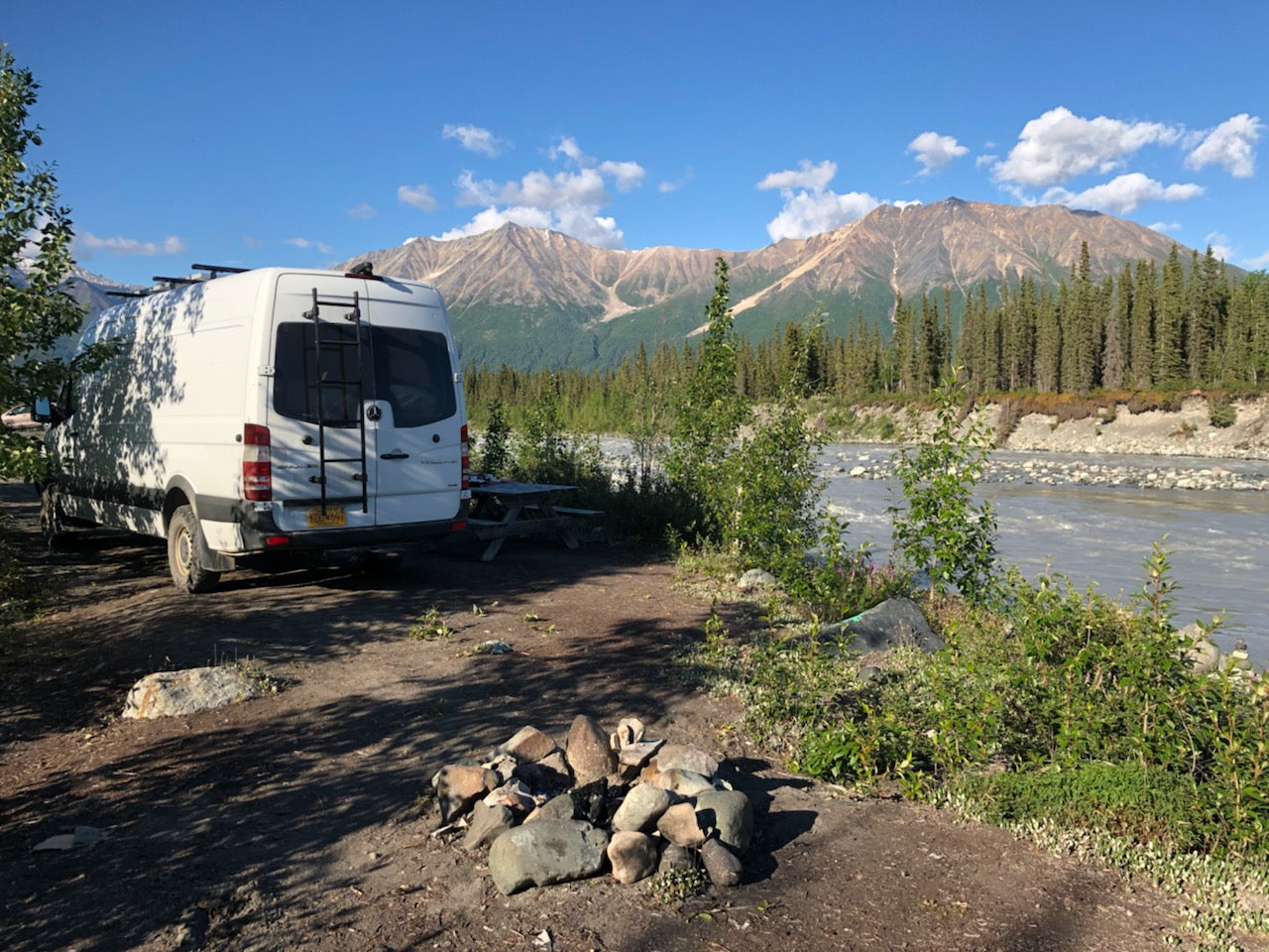 An RV rental parked riverside at a scenic campsite, with majestic mountains and a lush forest. Ideal for wilderness exploration.