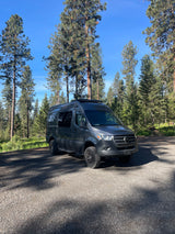 Gray Mercedes Sprinter camper van RV rental parked on gravel in a pine forest, ready for adventure.