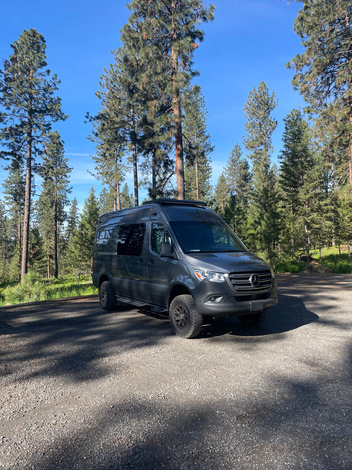 Gray Mercedes Sprinter camper van RV rental parked on gravel in a pine forest, ready for adventure.