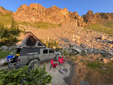 Jeep Gladiator RV rental: Overland rig with rooftop tent by a serene mountain lake. Campfire & chairs await adventure.