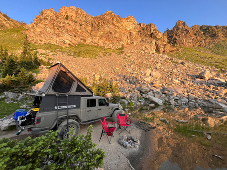Jeep Gladiator RV rental: Overland rig with rooftop tent by a serene mountain lake. Campfire & chairs await adventure.