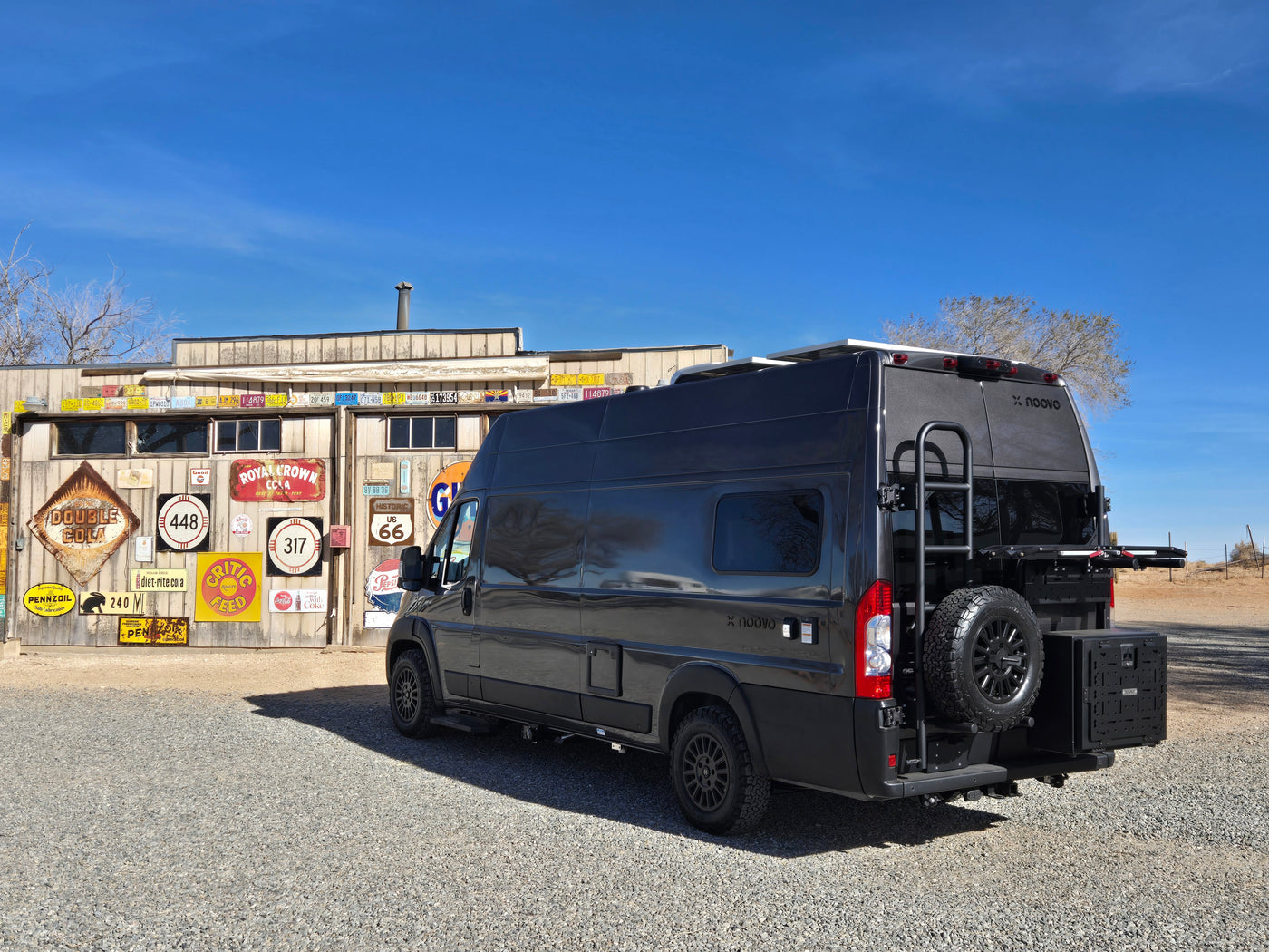 A dark grey Noovo rv rental van parked by a building adorned with vintage signs and license plates, ideal for road trips.