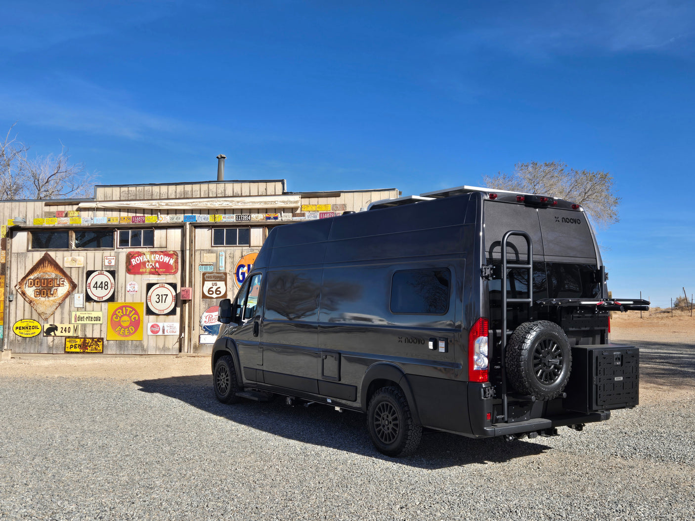 A dark grey Noovo rv rental van parked by a building adorned with vintage signs and license plates, ideal for road trips.