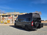 A dark grey Noovo rv rental van parked by a building adorned with vintage signs and license plates, ideal for road trips.