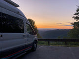 White RV rental parked at a mountain overlook during a colorful sunset, with rolling valleys below.