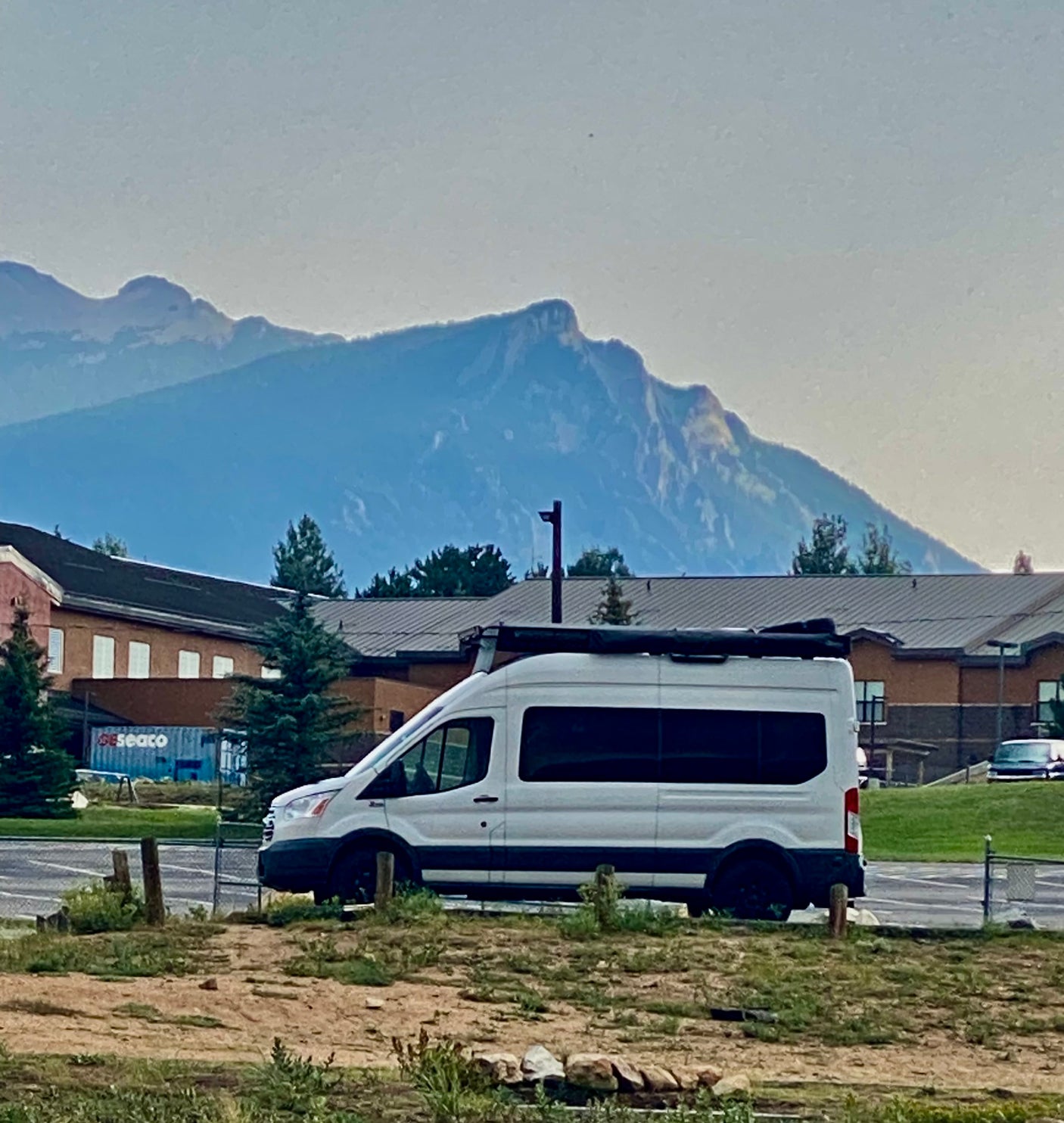 White Ford Transit RV rental van with awning, parked near mountains & buildings. Seaco container visible.