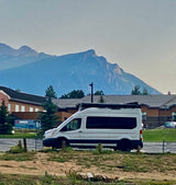 White Ford Transit RV rental van with awning, parked near mountains & buildings. Seaco container visible.