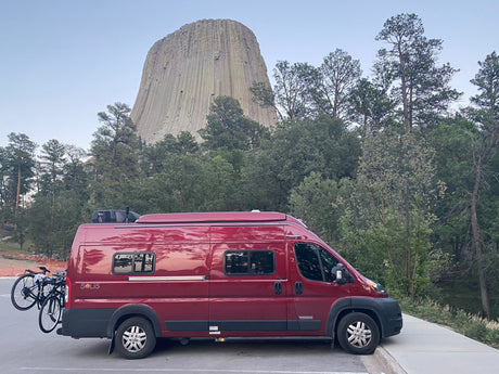 Red camper van rv rental with bikes parked at Devils Tower, surrounded by tall trees under a clear sky.