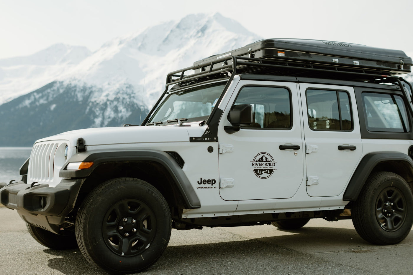 White Jeep Wrangler with a rooftop tent, available for RV rental from River Wild Alaska Camper Vans, set against snowy mountains. — photo 10