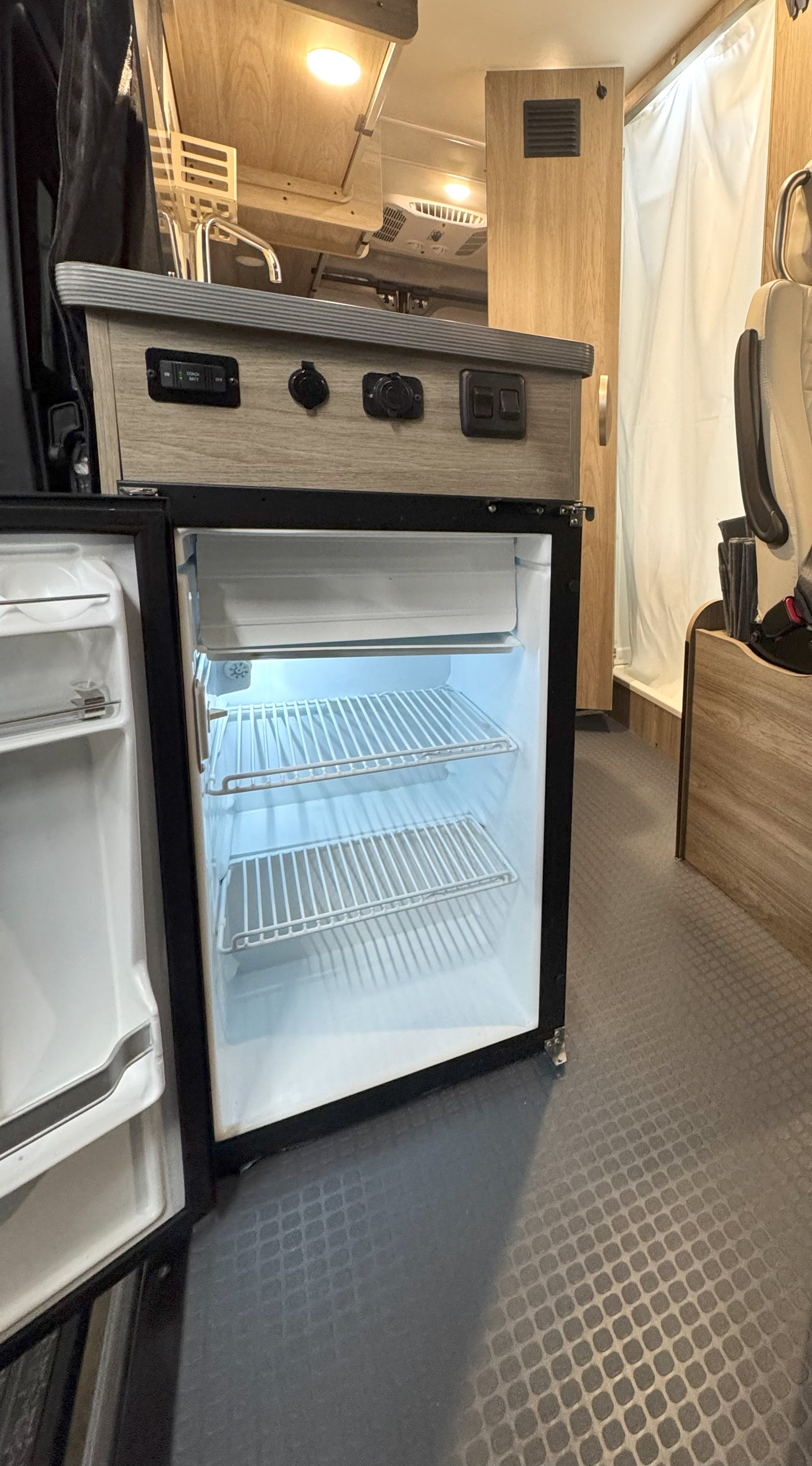 An open refrigerator and control panel on a wood-grain countertop inside an RV rental, highlighting the compact living space.