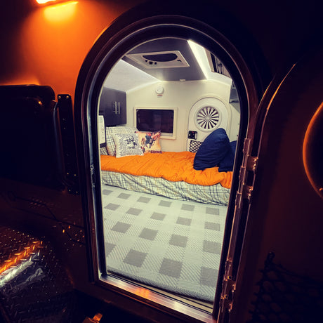 Inside this compact RV rental: a cozy bed with Goonies pillow, window, and warm lighting for adventures.