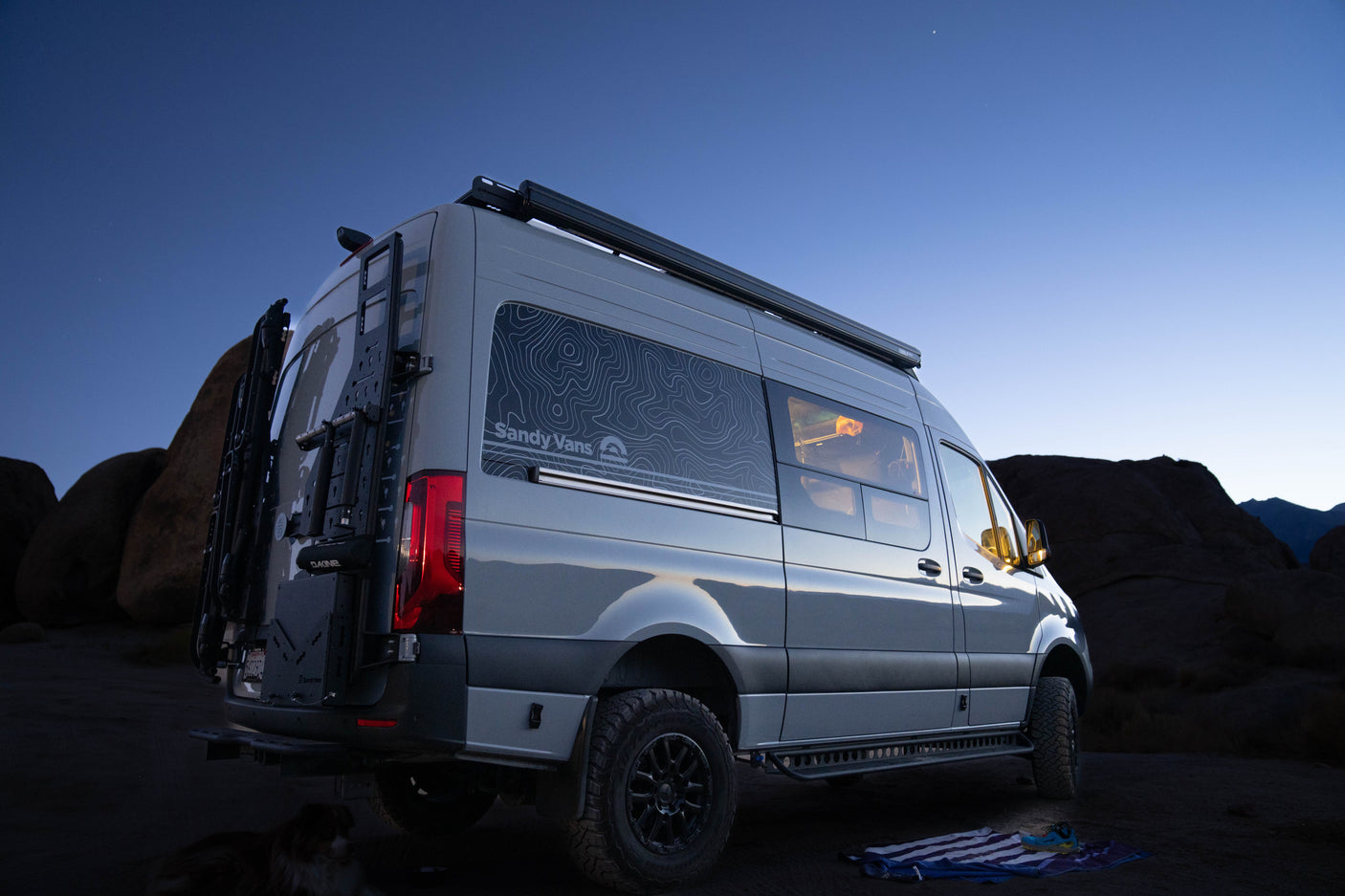 This grey Sandy Vans custom camper van rv rental is seen at dusk in a rocky landscape, equipped with off-road tires & gear.