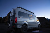 This grey Sandy Vans custom camper van rv rental is seen at dusk in a rocky landscape, equipped with off-road tires & gear.