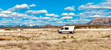 White RV rental parked in a vast dry field with distant snow-capped mountains under a blue, cloudy sky.