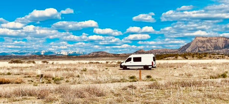 White RV rental parked in a vast dry field with distant snow-capped mountains under a blue, cloudy sky.