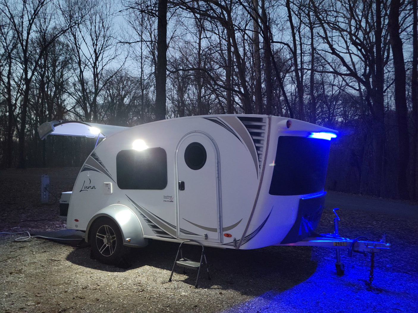 An illuminated white teardrop rv rental camper, with white and blue lights on, parked at a dark campsite surrounded by bare trees.