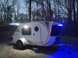 An illuminated white teardrop rv rental camper, with white and blue lights on, parked at a dark campsite surrounded by bare trees.