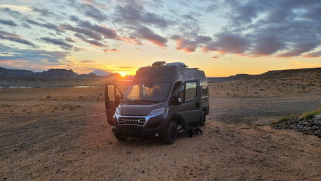 This RV rental camper van is parked on a dirt road at sunset, with warm light, clouds, mountains, and a lake in the background.