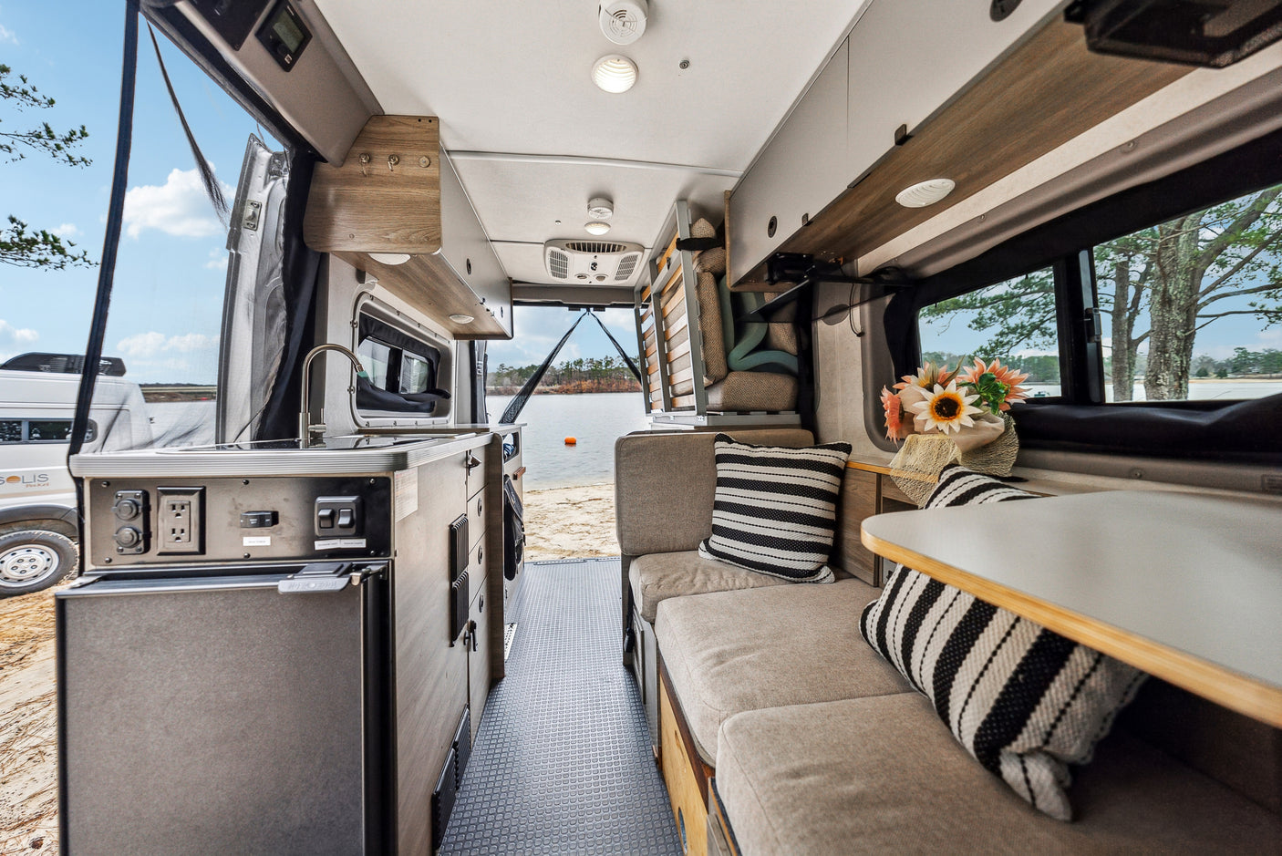Spacious RV rental interior features kitchenette & seating, opening to a serene lake view through the open back doors.