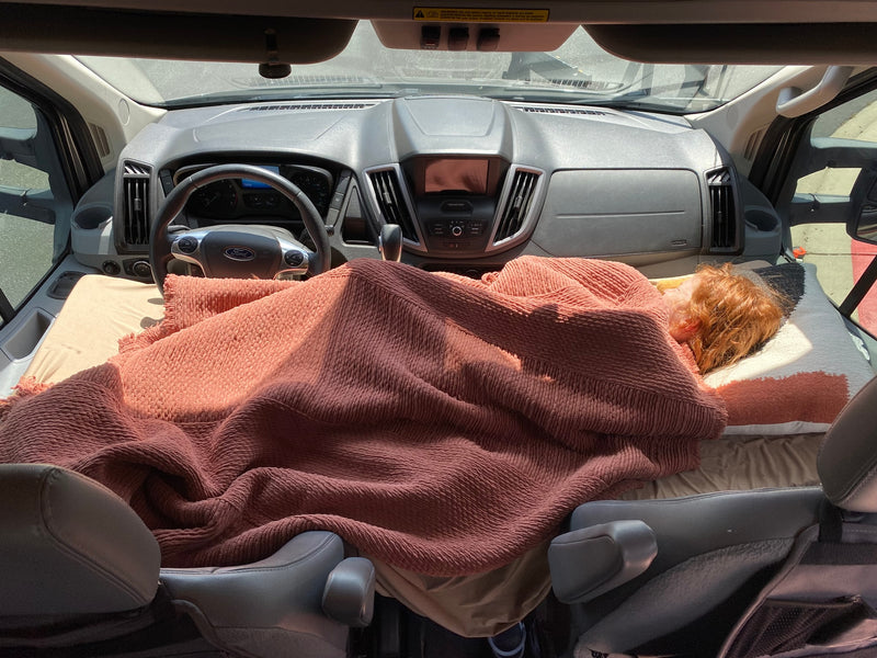 Person with red hair napping on a bed in the front of an **RV rental**, covered by a rust-colored blanket, inside a Ford camper van. — photo 12