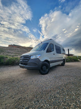A light-colored Mercedes Sprinter camper van, ideal for RV rental adventures, parked on a gravel road by a mesa.