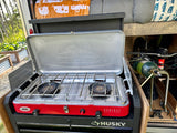 RV rental camping setup: A red Camp Chef Everest stove nested in Husky storage, with bike gear & propane for outdoor cooking.