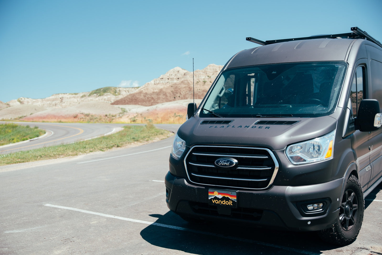 A dark gray Flatlander rv rental van is parked on asphalt, facing a scenic road and colorful badlands mountains under a blue sky.