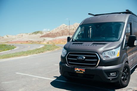 A dark gray Flatlander rv rental van is parked on asphalt, facing a scenic road and colorful badlands mountains under a blue sky.